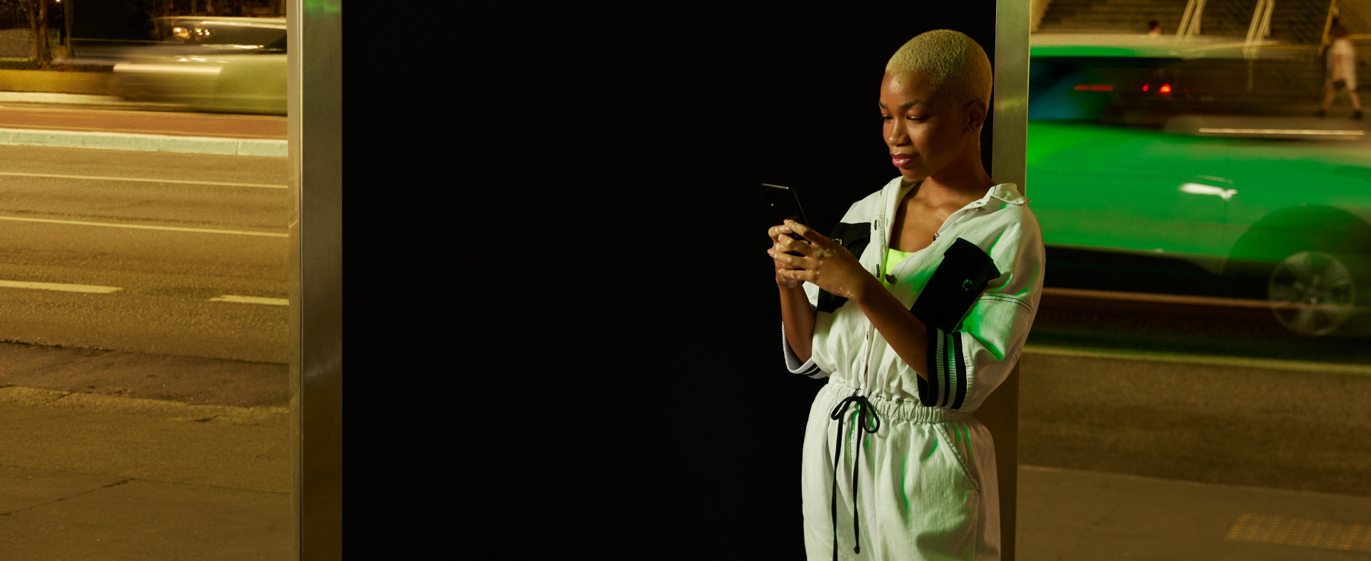 Uma mulher negra de cabelos raspados e descoloridos segura um smartphone com as duas mãos. Ela está em um ambiente urbano noturno e atrás dela passa um carro verde neon.
