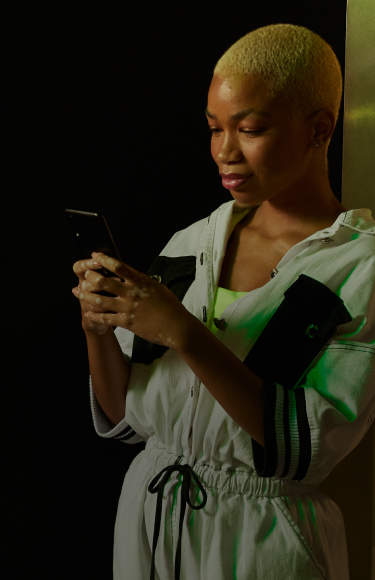 Uma mulher negra de cabelos raspados e descoloridos segura um smartphone com as duas mãos. Ela está em um ambiente urbano noturno e atrás dela passa um carro verde neon.