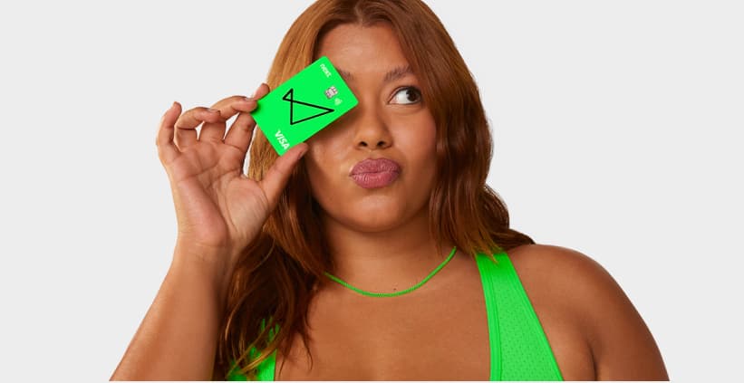 Uma mulher de pele morena e cabelos ondulados ruivos segura um cartão next na frente de um dos seus olhos. Ela usa uma blusa regata e colar de miçangas, ambos verdes neon do tom do cartão next.