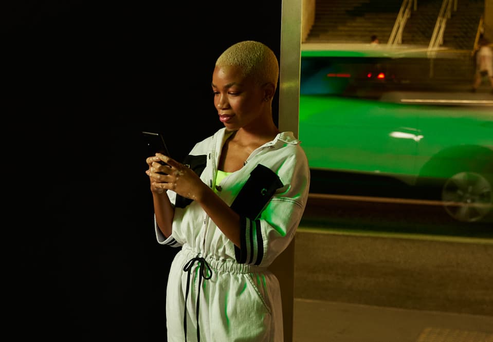 Uma mulher negra de cabelos raspados e descoloridos segura um smartphone com as duas mãos. Ela está em um ambiente urbano noturno e atrás dela passa um carro verde neon.