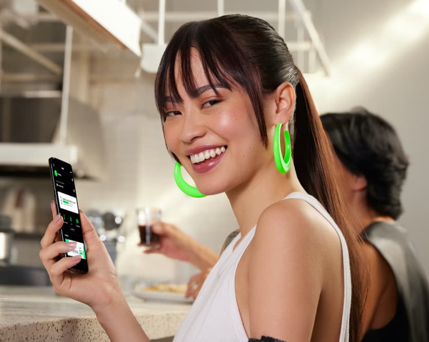 Duas pessoas estão sentadas em um balcão de uma padaria, tomando um café, suco de laranja e comendo um sanduíche. A pessoa da frente é uma mulher jovem de traços orientais, com o cabelo preso e brincos grandes verde neon. Ela sorri e segura um smartphone na mão com o aplicativo do next aberto nele.