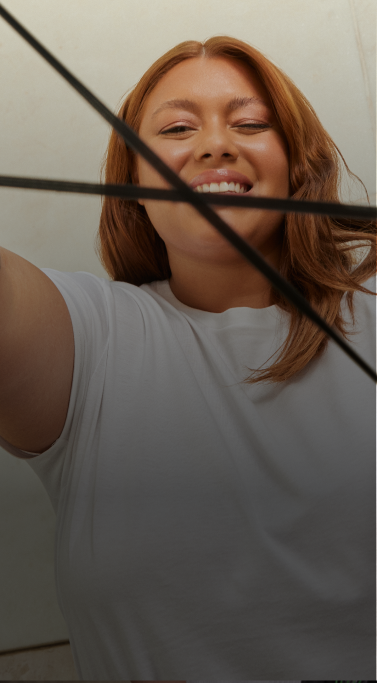 Uma mulher de pele morena e cabelos ondulados ruivos sorri enquanto olha para a câmera e faz o símbolo do logo do next com um elástico nas mãos, que lembra o símbolo do infinito.